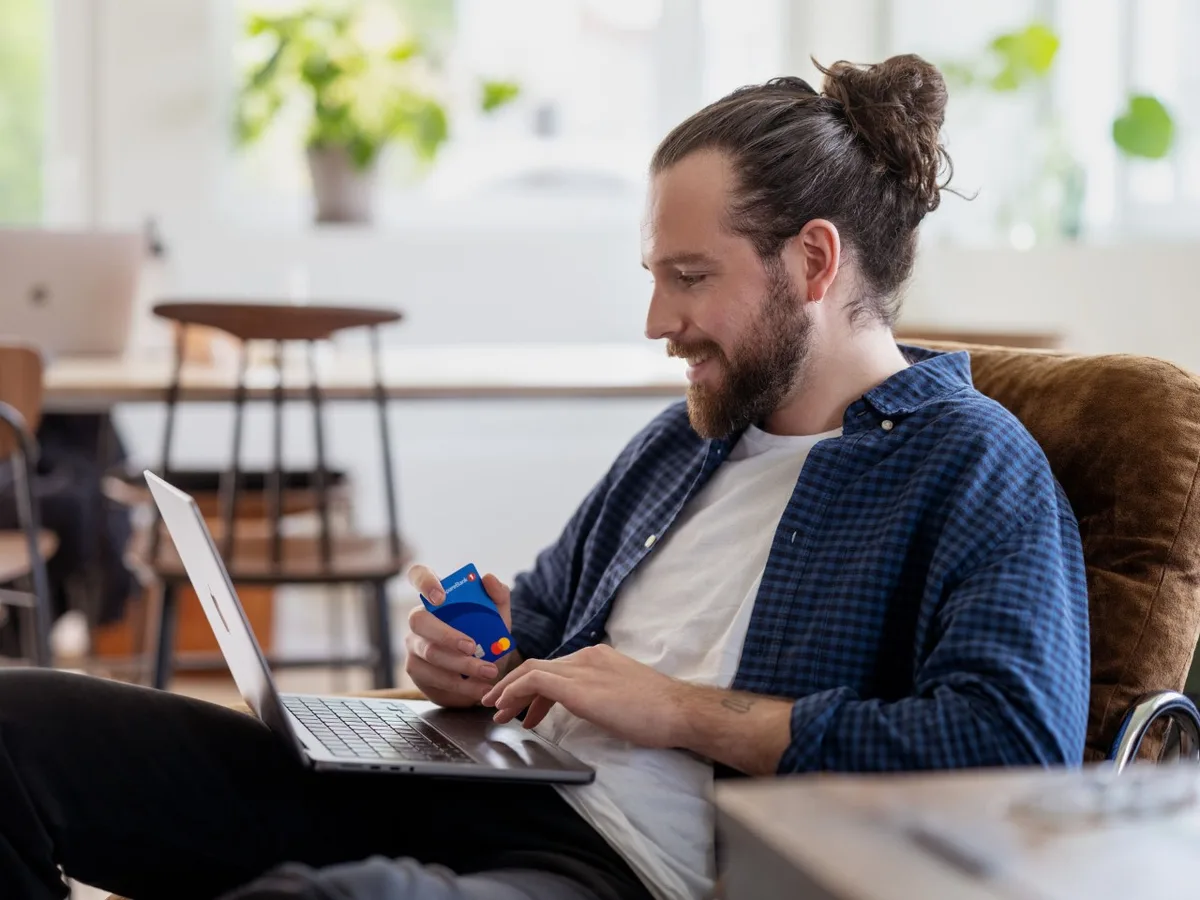 En mann sitter med laptop og et kredittkort i hånden