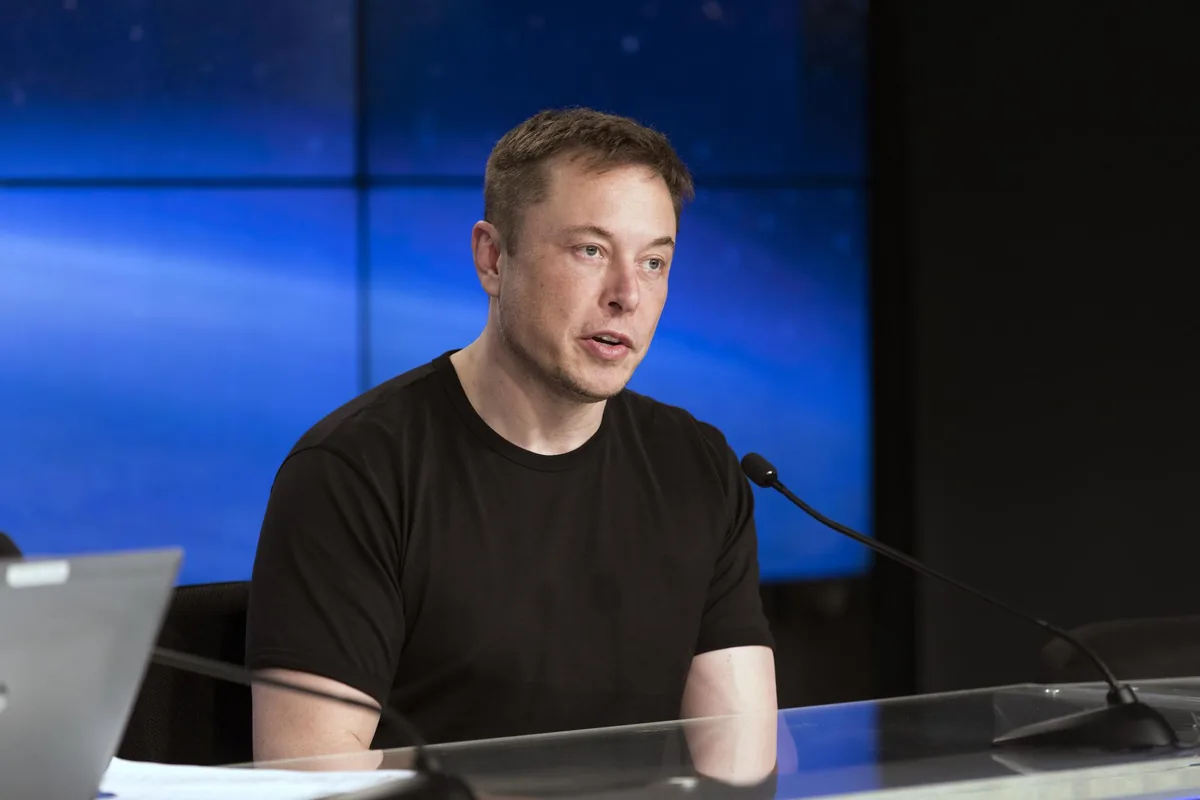 Elon Musk sitter ved et pressekonferansebord med mikrofon foran seg