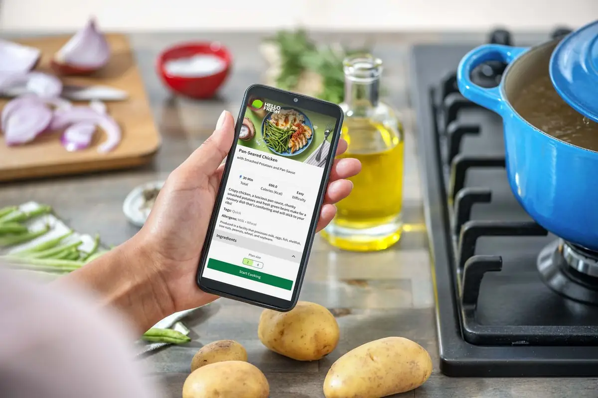 En hånd holder en mobiltelefon som viser en HelloFresh-oppskrift, foran en kjele på komfyren
