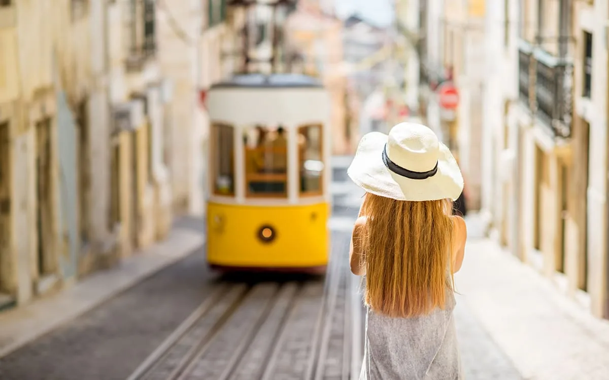 En turist med solhatt ser på en gul trikk i en smal gate i Lisboa